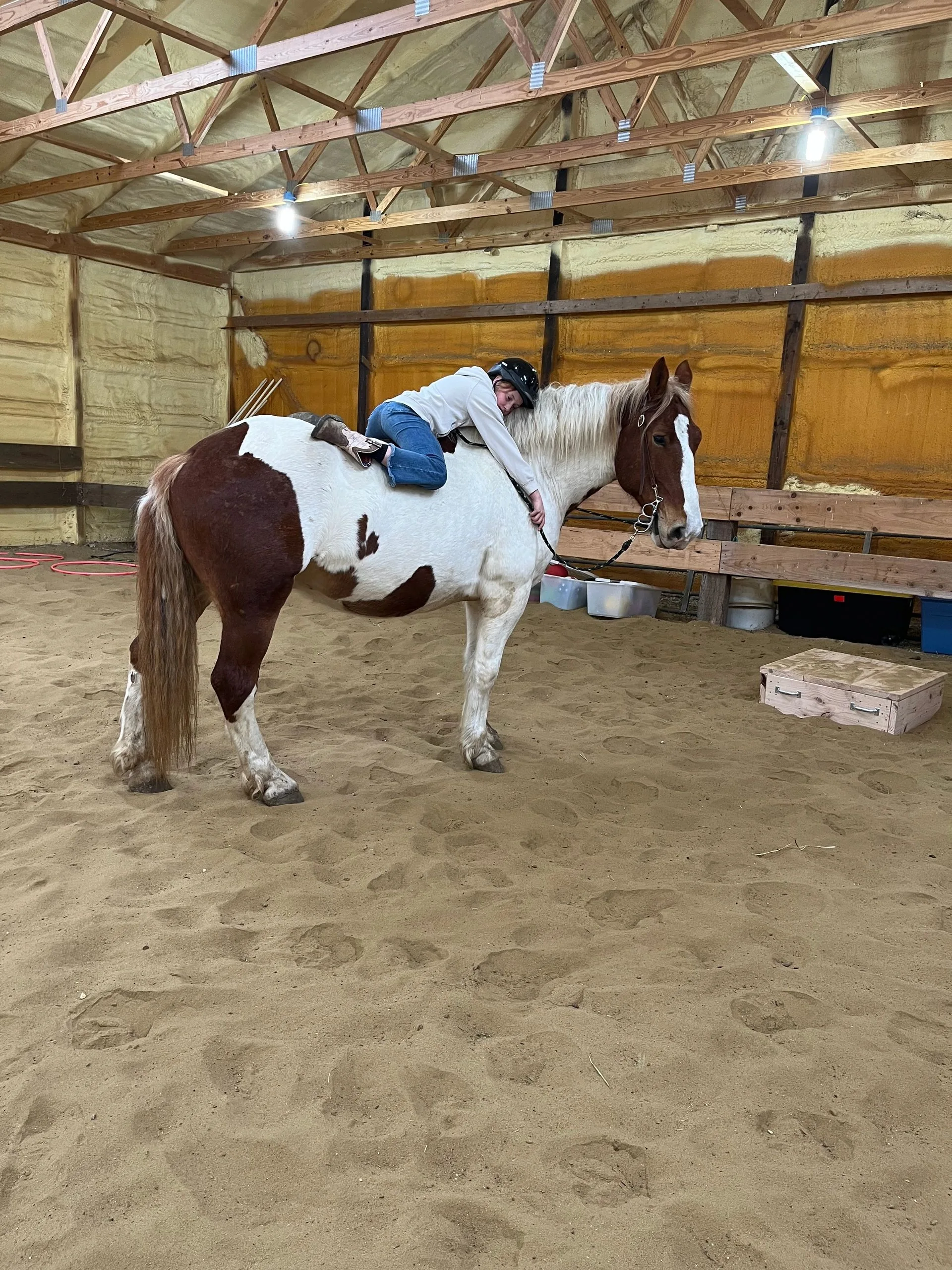 A helmeted rider leaning forward to hug a paint horse in the ranch arena